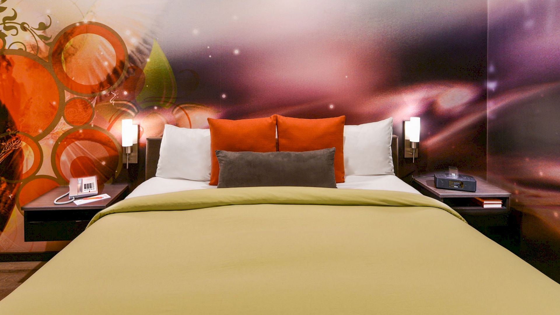 A modern hotel room bed with orange-and-white pillows, bedside lamps, and an abstract mural behind it.