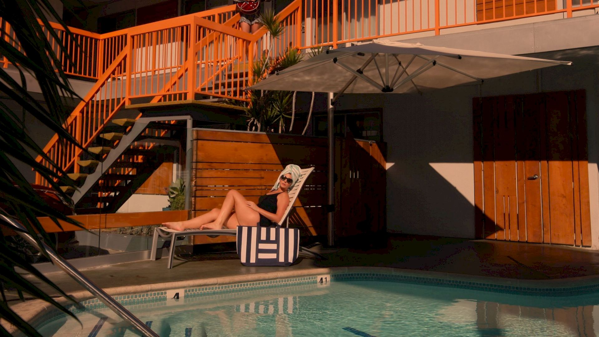 An outdoor pool area: a woman sunbathes on a chaise by the water, while a guitarist performs on an upper balcony above.